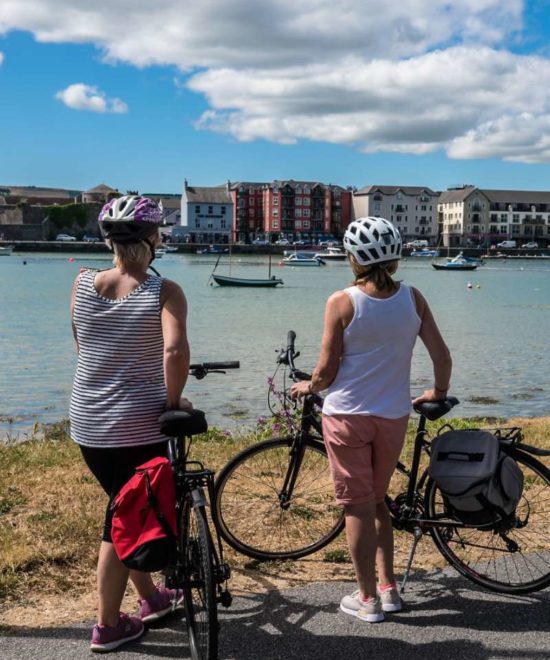 Two people wearing helmets stand with bicycles, part of a Copper Coast cycle tour, overlooking a scenic harbor. The water is calm with a few boats, and colorful buildings line the opposite shore. The sky is partly cloudy, suggesting a leisurely, sunny day perfect for cycling adventures.