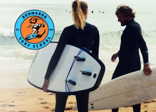 Two people in wetsuits stand on the sandy beach of the Copper Coast, surfboards by their sides as waves crash behind them. A logo in the top left corner reads "Bunmahon Surf School," featuring a surfing figure illustration.