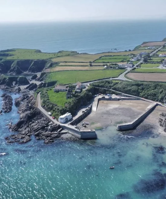 Aerial view of the scenic Dunabrattin Head on the Copper Coast, with rocky cliffs and turquoise waters. A small harbor with boats is nestled among green fields and winding roads. The sky is clear, and the horizon stretches into the distance.