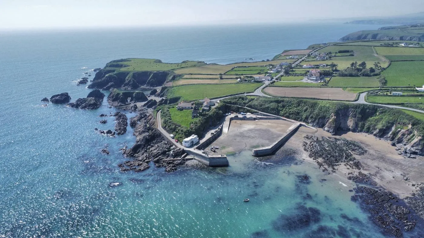 Aerial view of the scenic Dunabrattin Head on the Copper Coast, with rocky cliffs and turquoise waters. A small harbor with boats is nestled among green fields and winding roads. The sky is clear, and the horizon stretches into the distance.