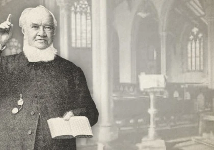 An older man with a beard, wearing glasses and holding an open book, gestures with one hand. He stands against a blurred backdrop of the Copper Coast church interior, where tall arches and stained glass windows echo stories reminiscent of David Alfred Doudney's heritage.