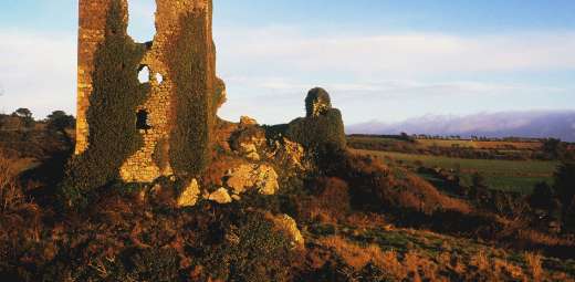 A partially ruined stone tower, reminiscent of Dunhill Castle, covered in ivy, sits on a grassy hill under a clear blue sky. The surrounding landscape along the Copper Coast features fields and distant hills, creating a peaceful rural scene.