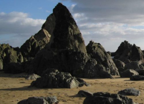 A rocky beach landscape at Garrarus Strand, with large jagged rocks protruding from the sand under a partly cloudy sky on the stunning Copper Coast. The light and shadows create a dramatic, contrasting effect.