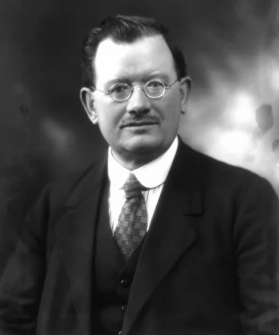 A black and white portrait of John Wheatley, the Labour MP, wearing round glasses, a suit, and a tie. He has short, combed-back hair and a mustache. The background is softly blurred.