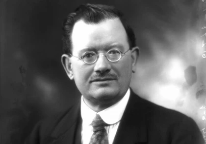 A black and white portrait of John Wheatley, a notable Labour MP, wearing round glasses and a suit with a tie. He has short, neatly styled hair and a mustache, gazing directly at the camera against a softly blurred background that echoes his place in history.