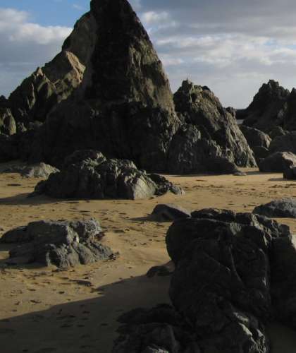 The Rocky beach scene at Kilfarrasy features large jagged rocks casting shadows on the sandy strand. A partly cloudy sky adds contrast to the dark rocks and golden sand.