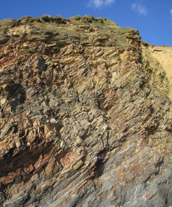 A rugged cliff face at Kilfarrasy Strand showcases layers of dark brown and gray rock formations, dotted with small patches of green vegetation at the top. Above this stunning coastal scenery, the sky remains clear and blue.