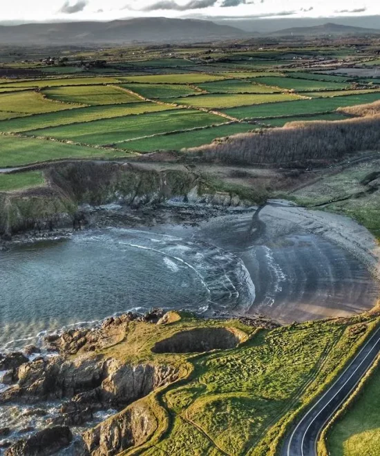 Aerial view of the scenic Copper Coast featuring Kilmurrin Cove's rocky cove and sandy beach. Surrounding the coast are lush green fields bordered by stone walls and a winding road. In the background, rolling hills stretch under a cloudy sky.