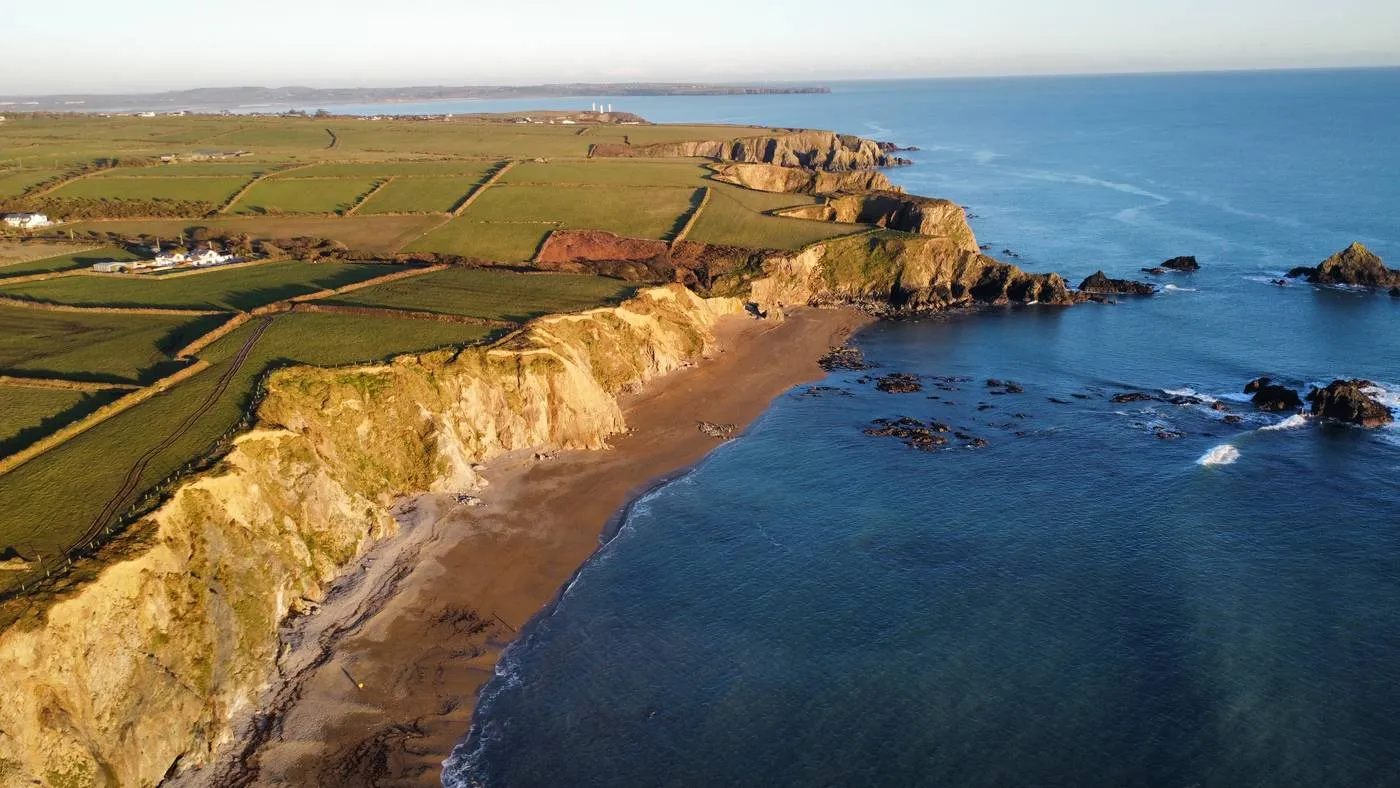 Aerial view of the rugged coastline along Garrarus Strand on the Copper Coast, with lush green fields atop cliffs, a sandy beach below, and rocky outcrops in the blue sea. More cliffs and open sea stretch across the horizon under a clear sky.