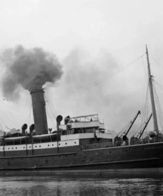 Black and white photo of a vintage steamship with a single smokestack emitting smoke, echoing the legacy of Copper Coast shipwrecks. The vessel's long deck is reflected in the calm harbor waters. The sky is overcast.