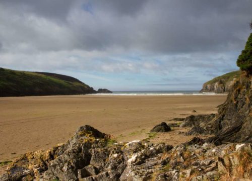 A serene beach scene at Stradbally Cove, with a sandy shore surrounded by rocky outcrops and green hills under a cloudy sky. The ocean is visible in the background, creating a calm and natural atmosphere.