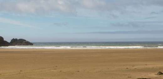 A wide, sandy beach stretches toward a calm sea under a partly cloudy sky. At Stradbally Cove, small waves gently roll onto the shore, with rocky outcrops visible in the distance on the left.