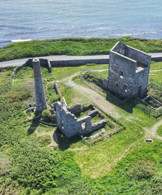 Aerial view of the Tankardstown stone ruin on a grassy coastline. The site features a chimney, partial walls, and a rectangular stone structure. A narrow road curves along the coastline, with the sea framing the picturesque background.