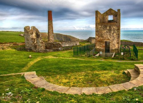 A coastal landscape featuring the historic stone ruins of Tankardstown, including a tall chimney and partially standing buildings. Green grass surrounds the scene, with the sea and a cloudy sky providing a dramatic backdrop.