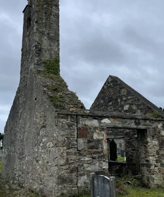 An old stone ruin in the Village of Kill, Ireland, with a tall chimney partially covered in ivy, stands against a cloudy sky. A gravestone with flowers in the foreground adds a somber tone to the scene, while grass and remnants of a building surround this historic site.