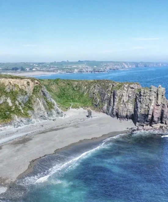 Aerial view of a rugged coastline at Tra na mBo, with tall cliffs, a sandy beach, and blue ocean waves. The foreground shows rocky formations jutting into the sea, while the background features a lush green landscape under a clear blue sky.