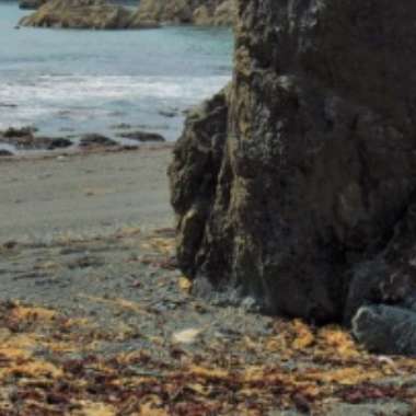 A rocky beach scene with a large rock formation in the foreground, reminiscent of Tra na mBo. The ocean stretches into the background, gentle waves meeting the sandy shore adorned with seaweed. Small distant rocks quietly protrude from the water beyond.