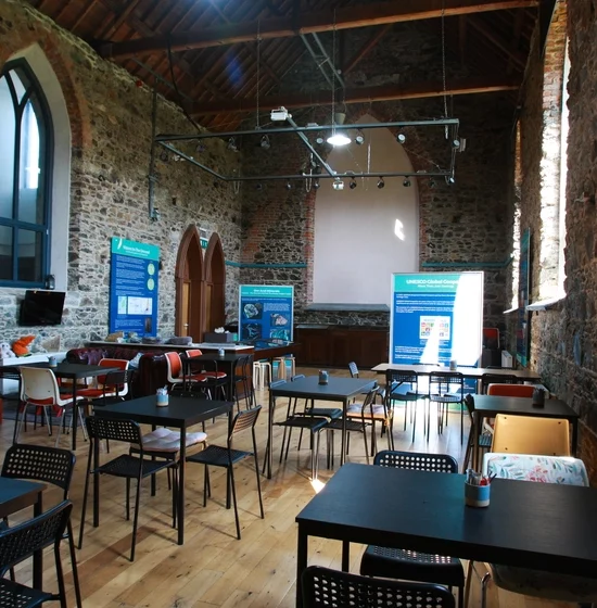 A rustic classroom interior at the Copper Coast Visitor Centre features exposed stone walls and a high wooden ceiling. Several black tables and mixed chairs are neatly arranged, while large arched windows let in natural light. Informational posters are prominently displayed at the front.