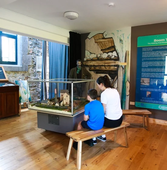 A woman and child sit on a bench at the Copper Coast Visitor Centre, observing a historical diorama. Behind them, an exhibit with informational posters and artifacts is displayed. The space features wooden floors, stone walls, and large windows that offer ample natural light.