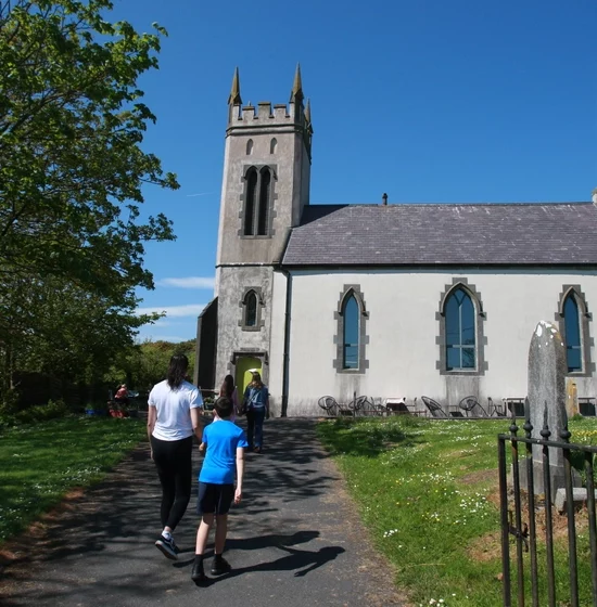 A group of people stroll along a path toward a small white chapel with a tall bell tower and arched windows. The sky is clear and blue, mirroring the serenity found around Copper Coast Visitor Centre. Trees and gravestones line either side of the path, whispering tales of time past.