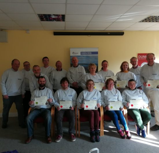 A group of people from the Copper Coast Network are standing and sitting indoors, proudly holding certificates and smiling at the camera. Clad in matching gray shirts, they gather in a room adorned with a seascape poster and a small red sign in the background.