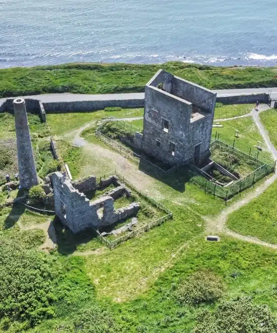 Aerial view of old mining ruins on a grassy cliff within the Copper Coast UNESCO Geopark. The site features a tall chimney and partially collapsed stone structure, with a winding path encircling the area. The shimmering ocean enhances the dramatic backdrop.