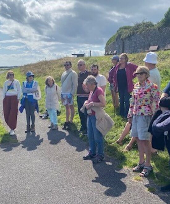 A group of people stand and sit along a seaside path on a sunny day, some with dogs. They are gathered near green grass and a stone wall, looking out towards the water. Some are dressed in casual summer clothing, with a mix of colors.