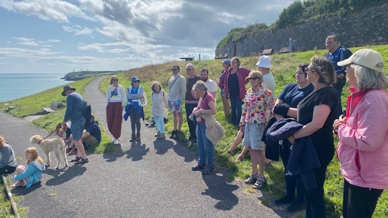 A group of people stand and sit along a seaside path on a sunny day, some with dogs. They are gathered near green grass and a stone wall, looking out towards the water. Some are dressed in casual summer clothing, with a mix of colors.