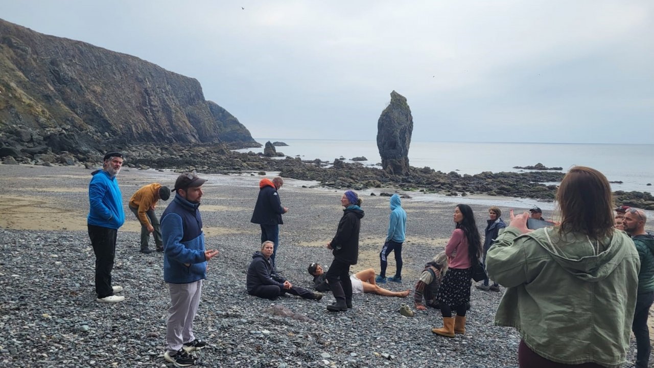 A group of people stands on a rocky beach near cliffs and a tall sea stack. Some are standing, while others are sitting on the pebbles. The cloudy sky creates a muted atmosphere.