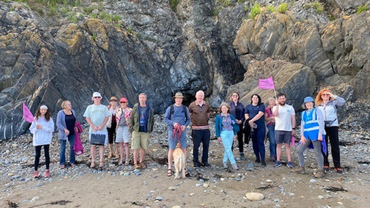 A diverse group of people stand on a rocky beach holding pink flags. They are dressed casually, with some wearing hats and sunglasses. A dog is present among the group. The background features large rock formations.