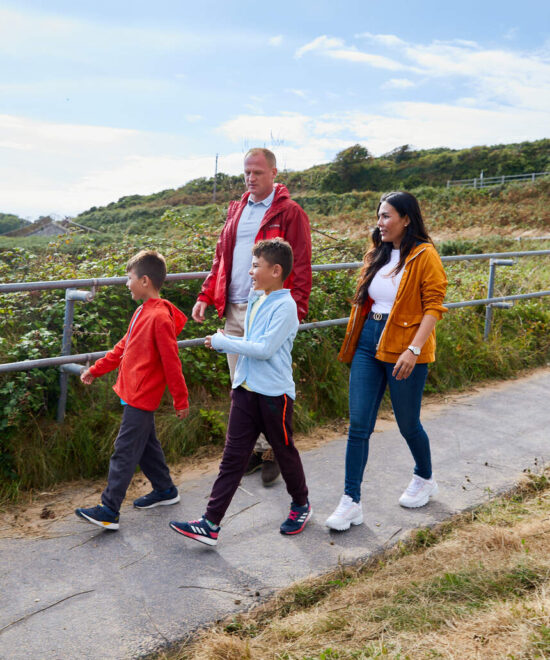 A family of four, including two adults and two children, strolls along a paved path by a grassy hillside under a partly cloudy sky. With places to stay nearby, one adult wears a red jacket, the other yellow. The children are ahead in matching red tops.